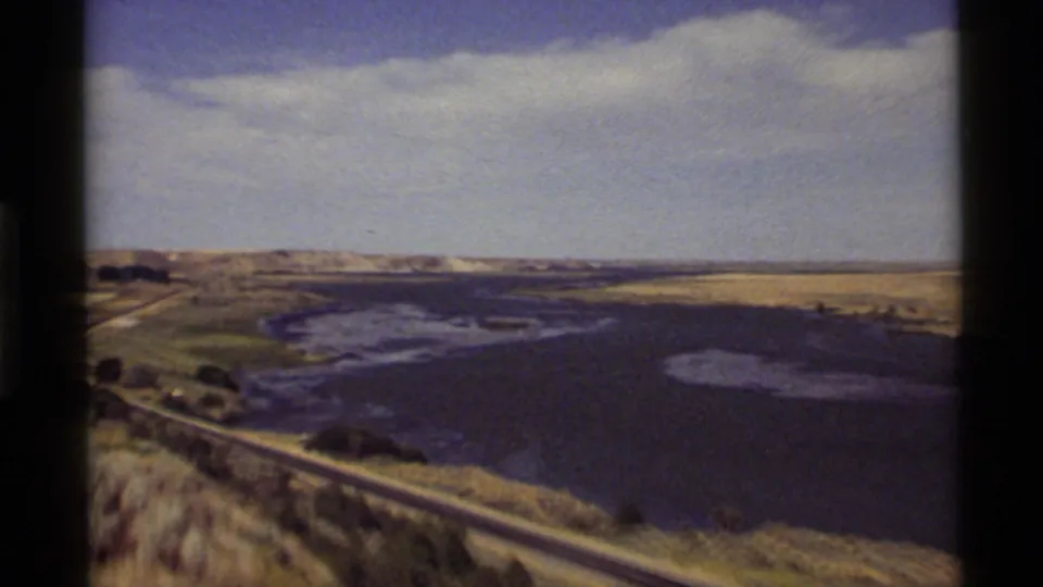 1972 Montana 8mm frame showing a broad water surface with pale shallows, roadside foreground, open land, and low distant hills.
