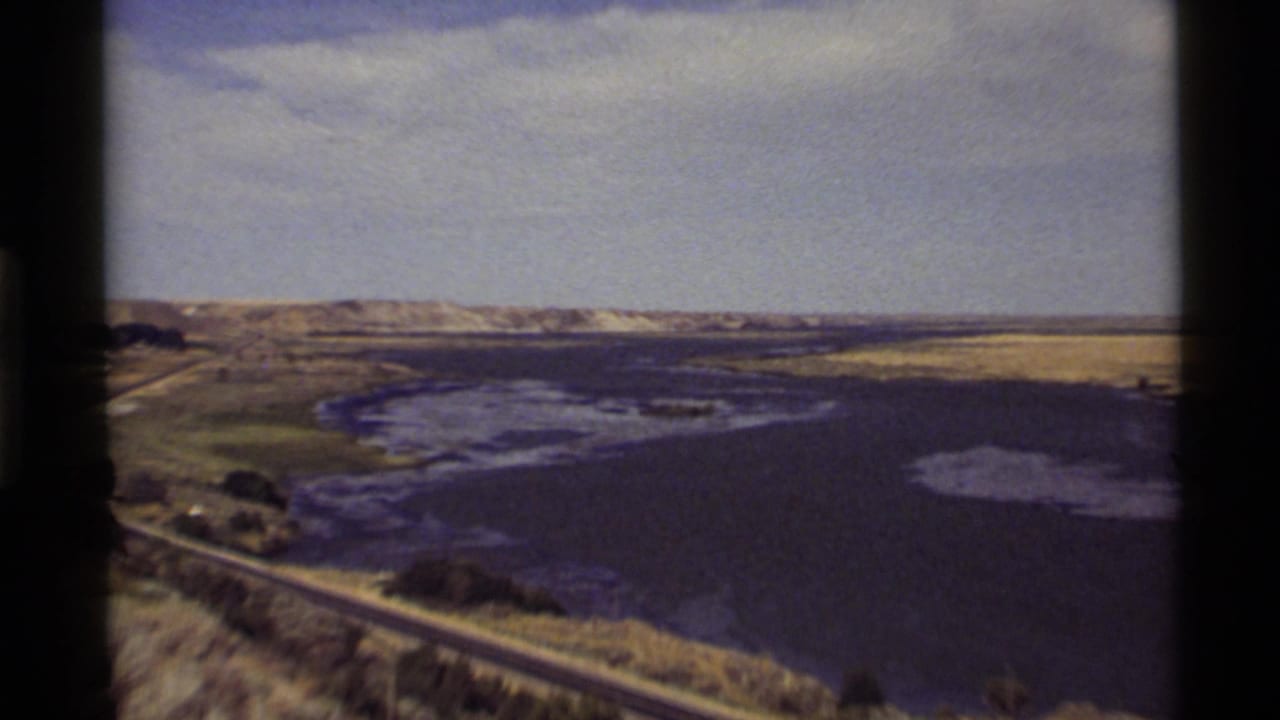 1972 8mm view of Holland Lake, Montana, with water, shoreline road, sparse greenery, distant hills, and pale sky.