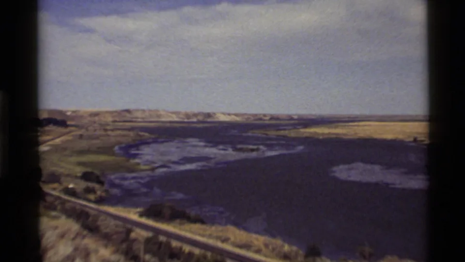 1972 8mm view of Holland Lake, Montana, with water, shoreline road, sparse greenery, distant hills, and pale sky.