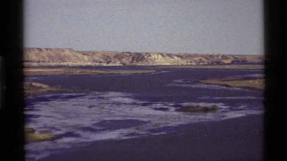 1972 8mm frame of Holland Lake with water in the foreground, pale hills across the horizon, and a narrow strip of shore.