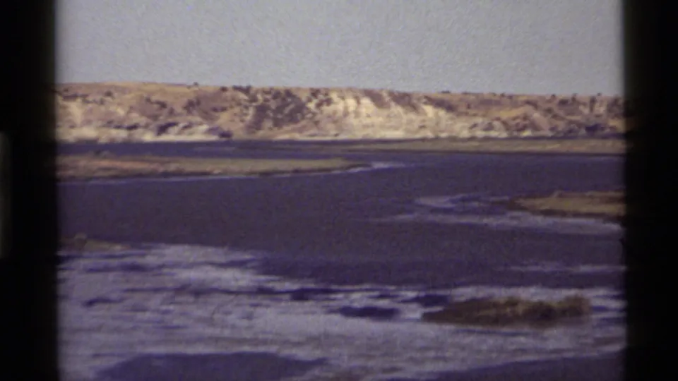 1972 Holland Lake, Montana frame showing calm water, marshy shallows, and a distant sunlit hillside on 8mm film.