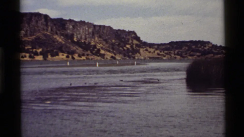 Holland Lake home-movie frame with ducks near the lower waterline, distant buoys, reeds, and a rugged Montana hillside.