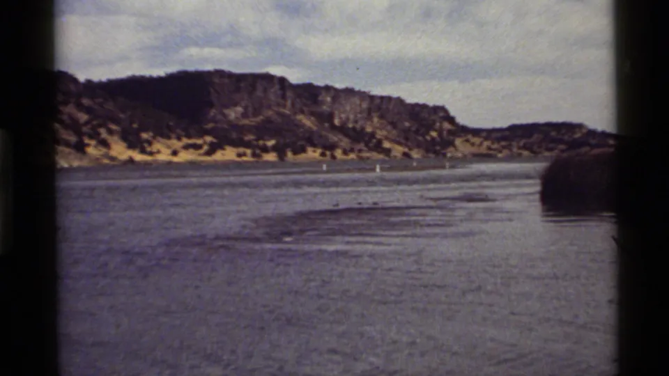 Final 1972 Holland Lake frame with rippled water in the foreground, buoy markers, dark reeds, and a rocky Montana hillside.