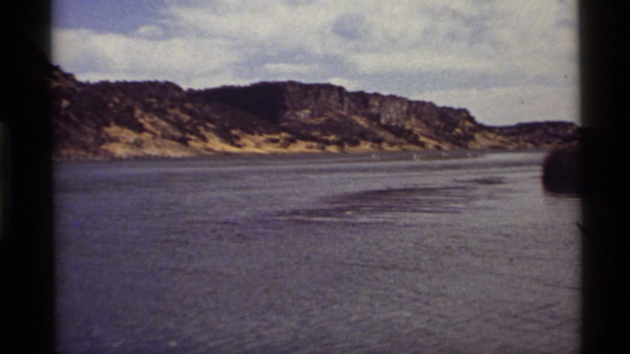 1972 8mm view of Holland Lake, Montana, with calm water, distant rocky ridges, sparse shoreline vegetation, and dark film-frame edges.