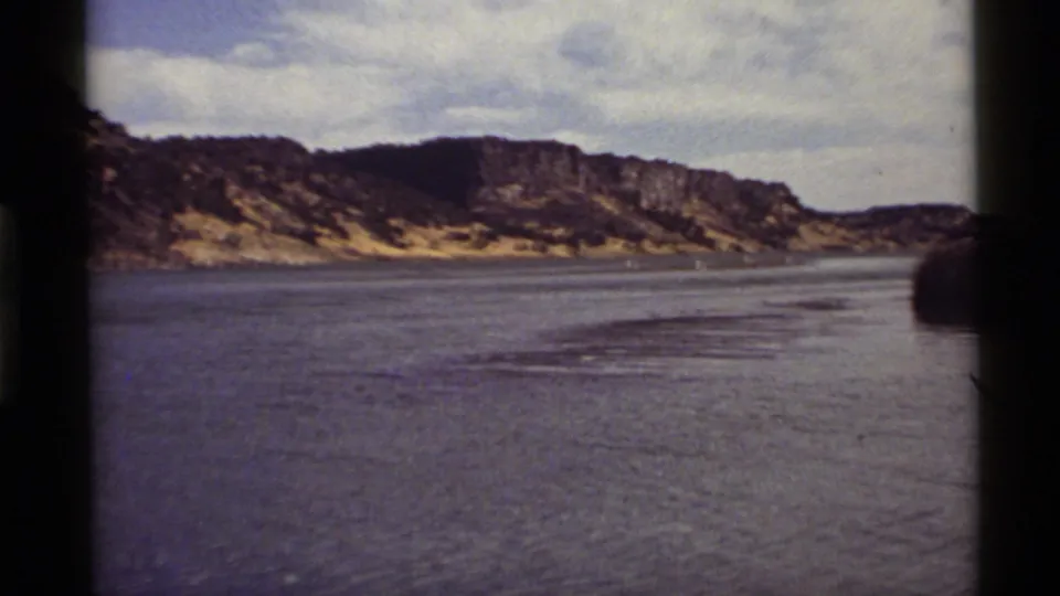 1972 8mm view of Holland Lake, Montana, with calm water, distant rocky ridges, sparse shoreline vegetation, and dark film-frame edges.