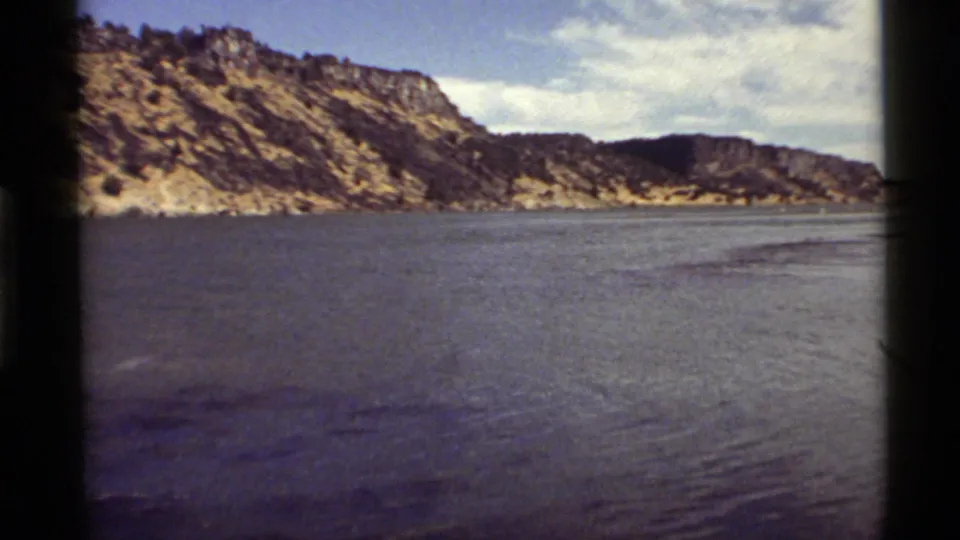 1972 home-movie frame of Holland Lake with textured water, rugged Montana ridges, sparse trees, and soft 8mm color.