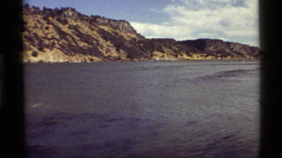 Holland Lake, Montana, shown in a soft 8mm frame with broad water, rocky hills, sparse vegetation, and partly cloudy sky.