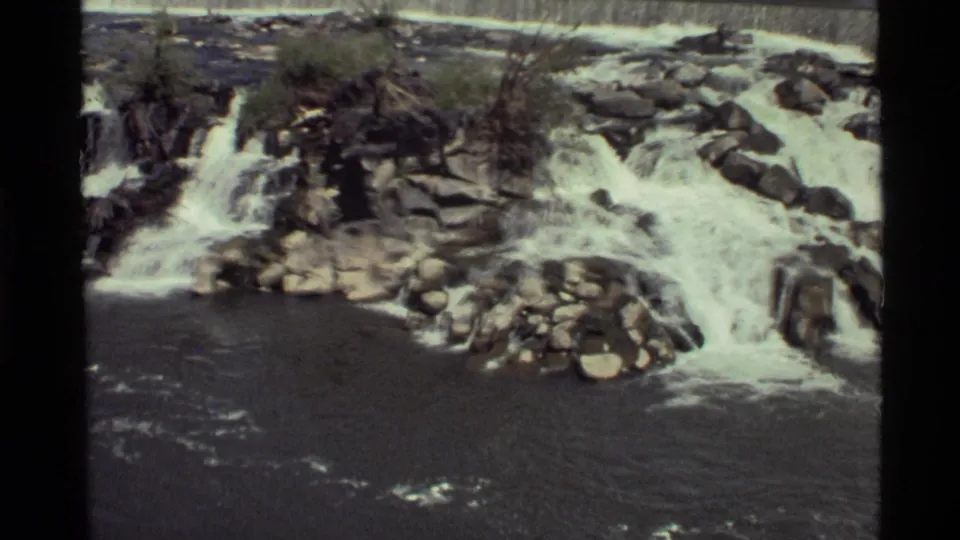 1972 8mm view of water spreading across rocky shelves at Holland Lake, with boulders and darker river water below.