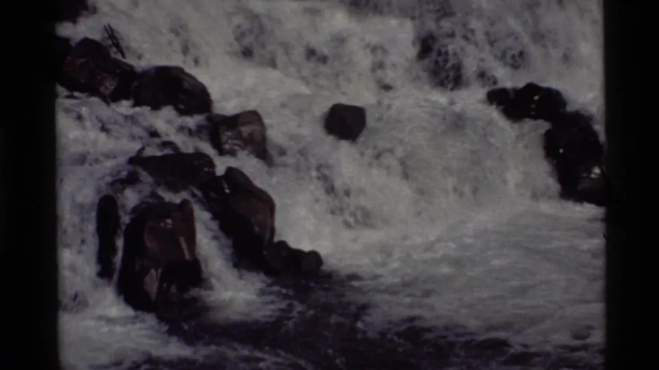 Foamy water courses through dark rocks at Holland Lake, Montana, in a grainy 1972 8mm home-movie frame.