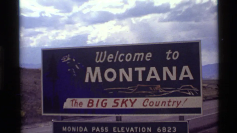 1972 8mm frame of the Welcome to Montana sign at Monida Pass, with open hills and bright clouds behind it.