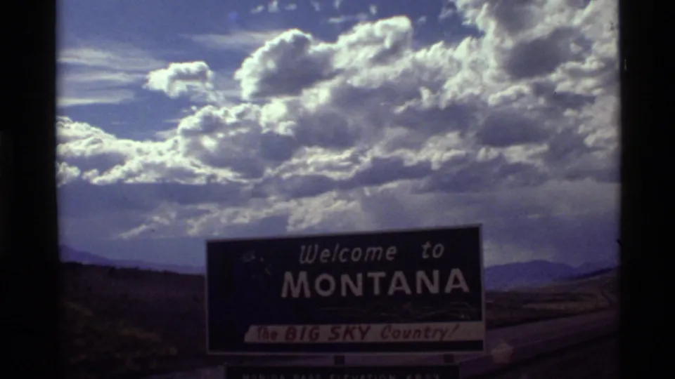 1972 home-movie frame showing a Montana welcome sign along the road, backed by distant hills and a cloud-heavy sky.