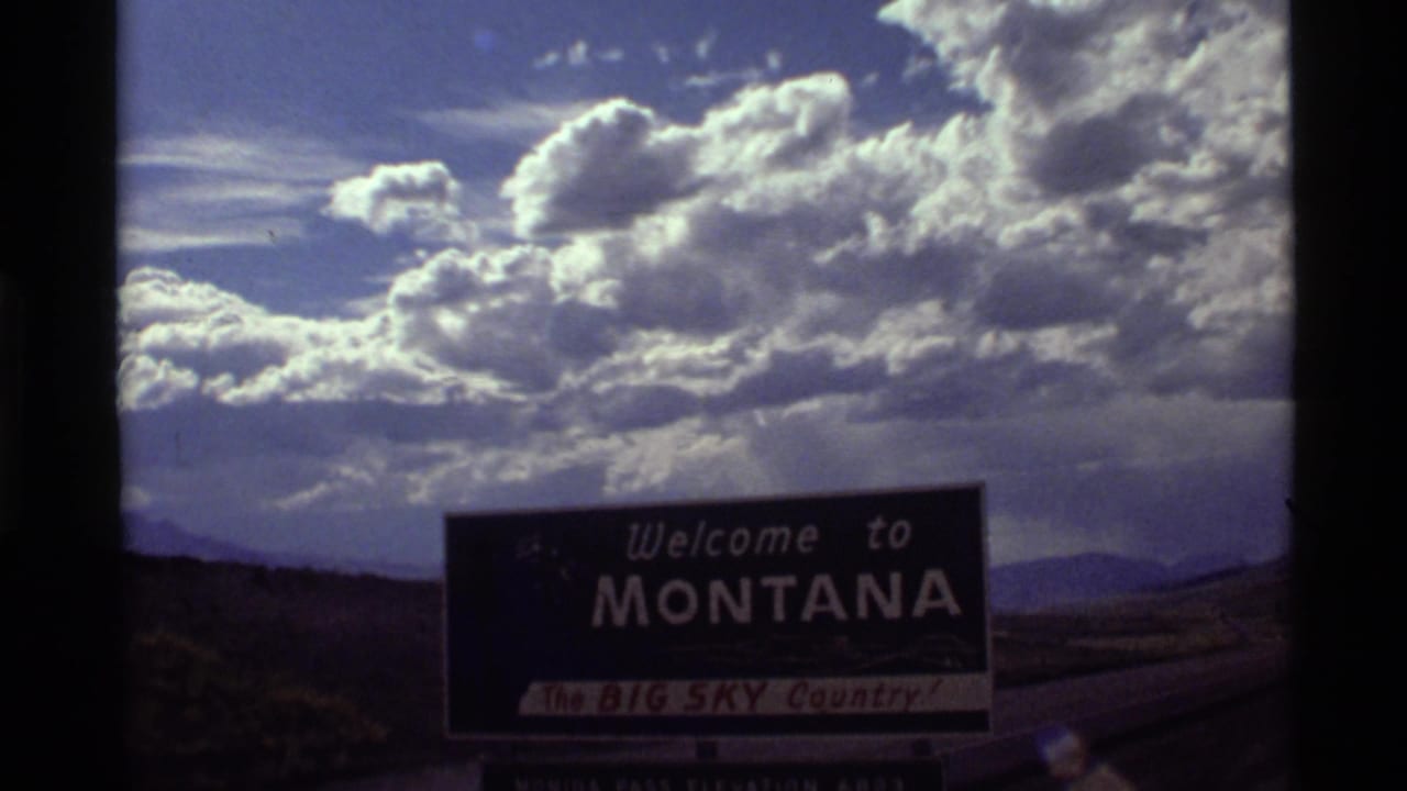 Montana welcome sign below a dramatic cloudy sky with distant hills.