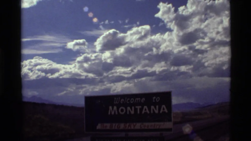 1972 Montana home-movie frame with the welcome sign low in the composition and bright cloud formations dominating the sky.