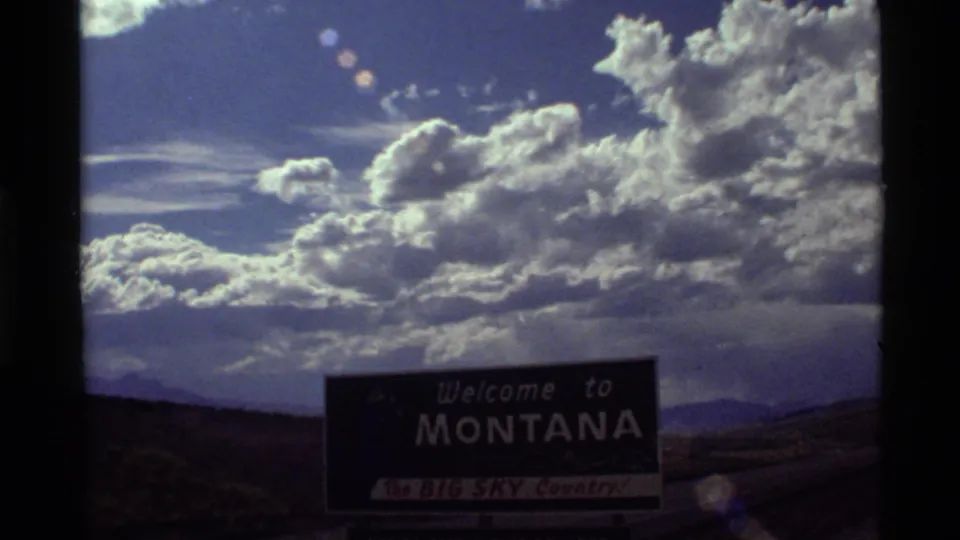 1972 8mm frame of the Montana welcome sign near the lower edge, with mountains and a luminous cloudscape above.