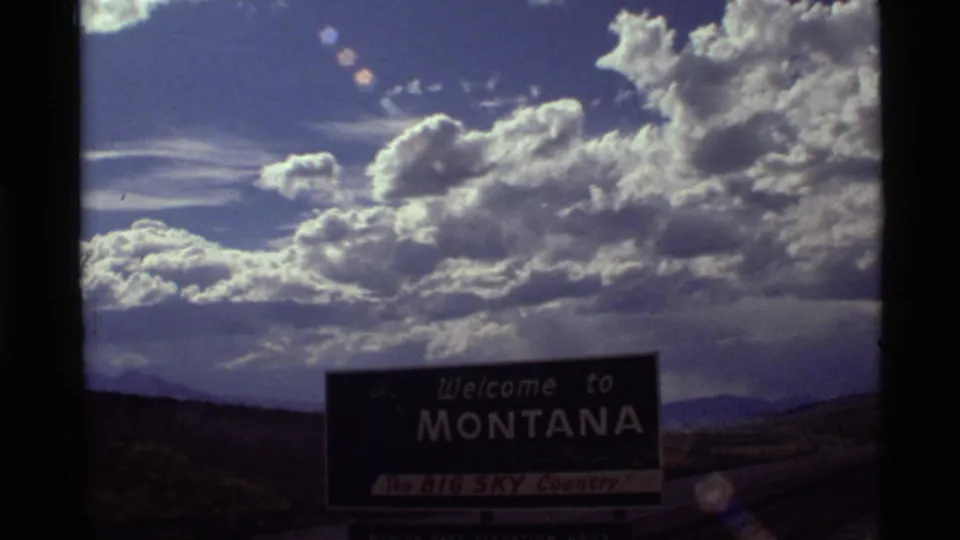 Final 1972 Montana road-trip still showing the welcome sign, distant mountains, lens flare, and dense sunlit clouds.