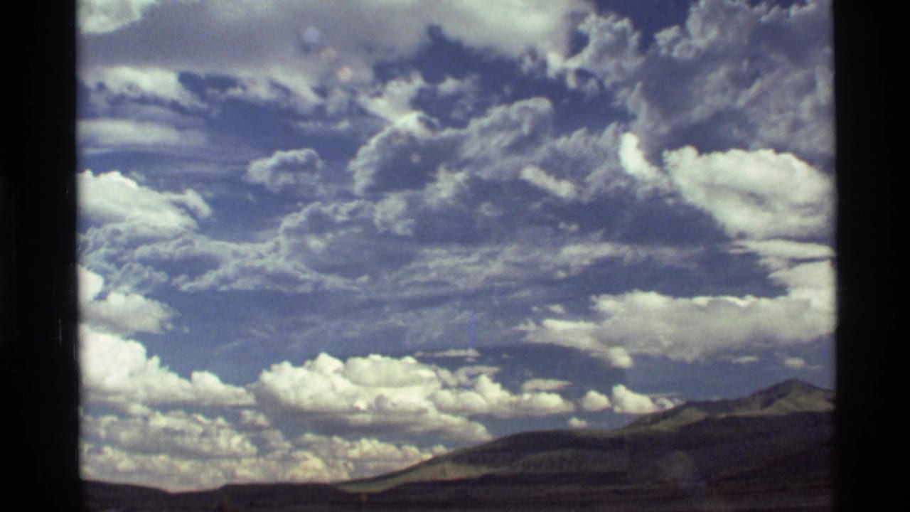 1972 8mm home-movie view near Holland Lake, Montana, showing bright clouds above a distant mountain range through vehicle-window framing.