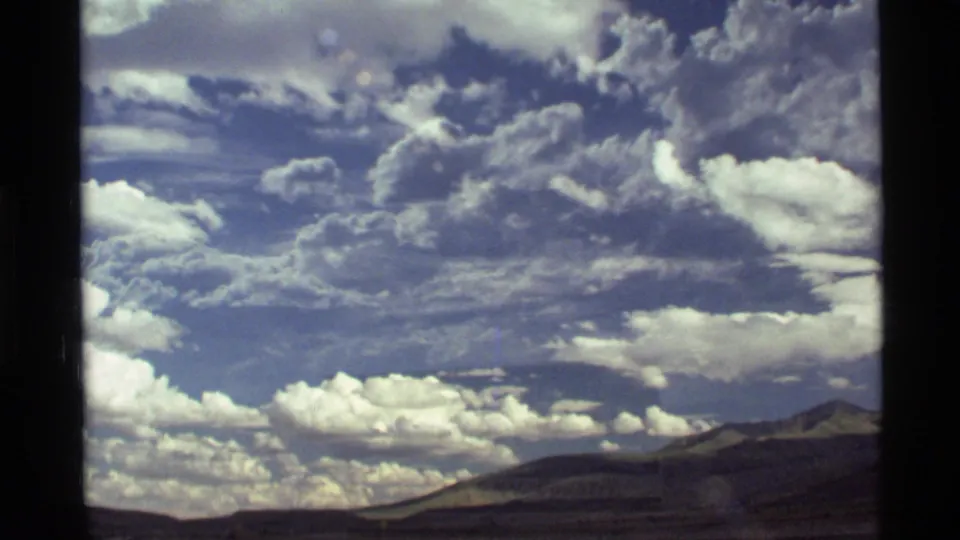 1972 8mm home-movie view near Holland Lake, Montana, showing bright clouds above a distant mountain range through vehicle-window framing.