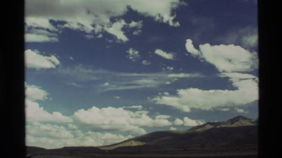 8mm home-movie frame from the Holland Lake, Montana clip showing bright clouds, open sky, and distant mountain slopes.