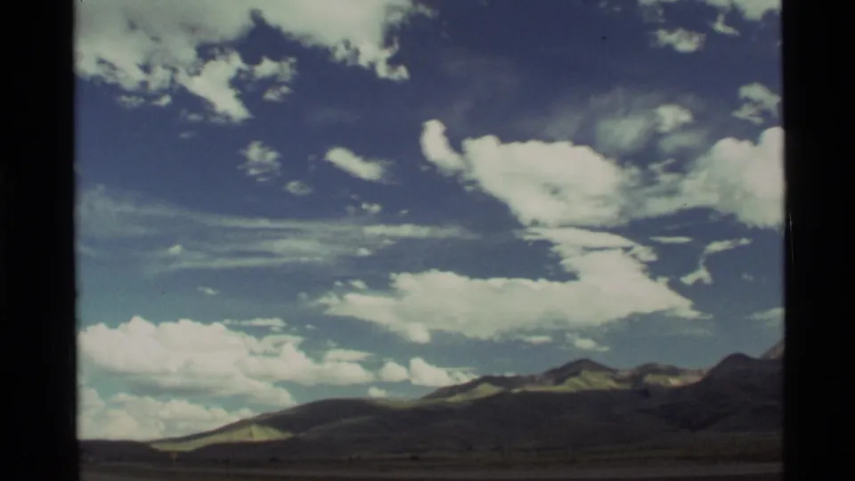 1972 8mm Montana landscape frame with large white clouds, dark mountain ridges, and road-trip window framing.