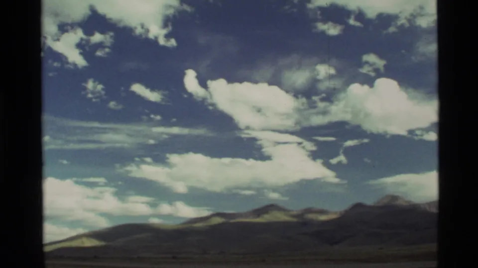 1972 8mm Montana landscape view with distant mountains, open plain, and bright cumulus clouds across a deep blue sky.