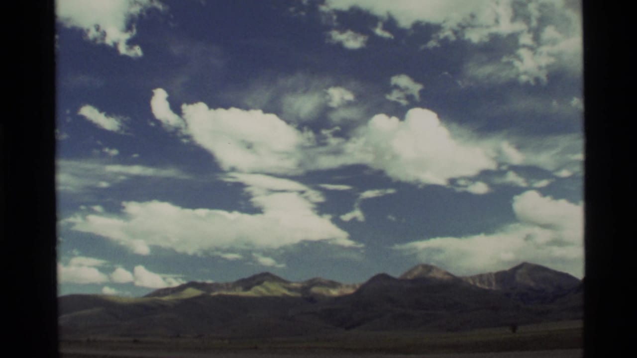 Vintage 8mm frame of the Holland Lake, Montana area showing a low mountain horizon, open land, and broken white clouds.