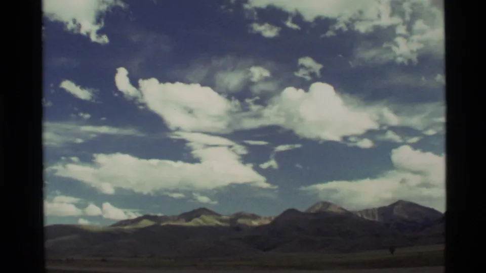 Vintage 8mm frame of the Holland Lake, Montana area showing a low mountain horizon, open land, and broken white clouds.