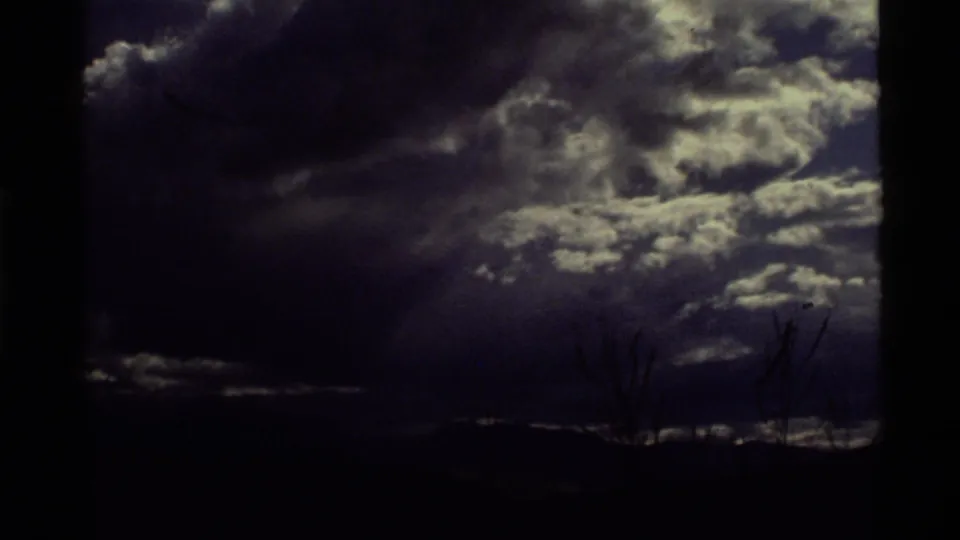 1972 8mm frame from Holland Lake, Montana, showing dark clouds, bright cloud breaks, mountain silhouettes, and sparse foreground branches.