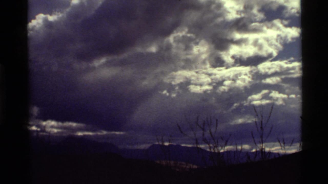 1972 8mm view near Holland Lake, Montana, with sunlit clouds, rays through overcast sky, distant mountains, and thin foreground branches.