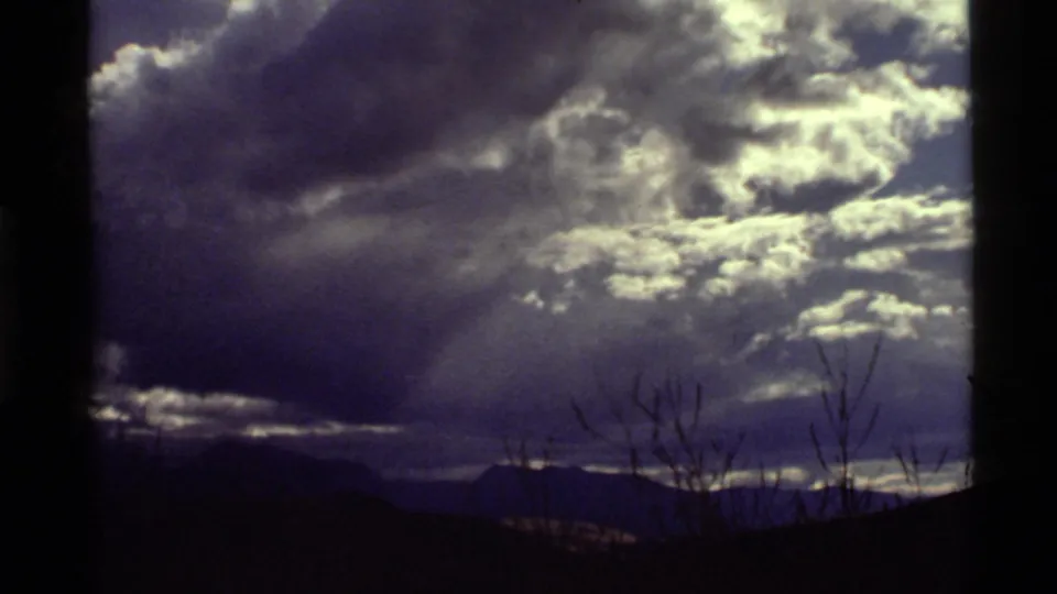 1972 8mm view near Holland Lake, Montana, with sunlit clouds, rays through overcast sky, distant mountains, and thin foreground branches.