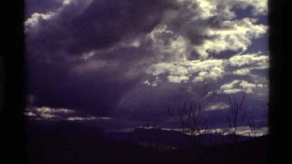 1972 Holland Lake, Montana frame showing layered clouds, bright sky openings, a dark ridge line, and silhouetted dry grasses or branches.