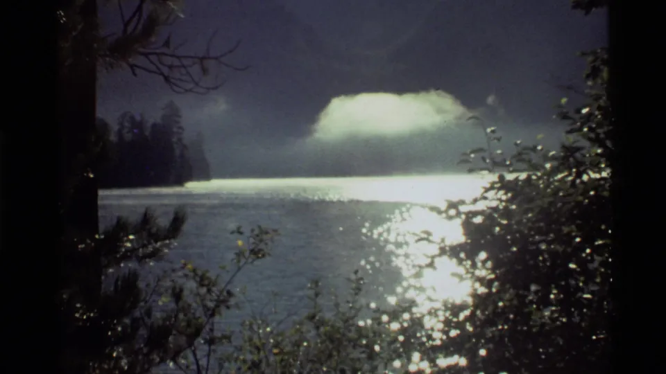 Final 1972 Holland Lake frame showing reflected sunset light on the water, a bright low cloud, and dark forest shapes at the shore.