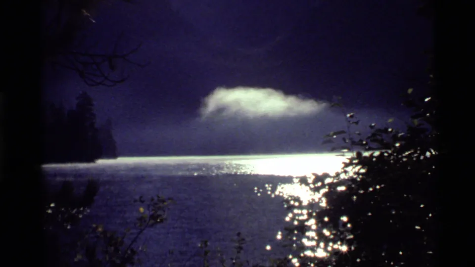 1972 Holland Lake, Montana 8mm frame showing a low illuminated cloud over dark water, with tree silhouettes and foreground leaves framing the view.