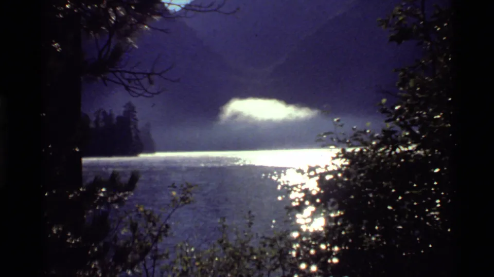 1972 Holland Lake, Montana frame with silhouetted trees at left and right, a bright reflective lake surface, and a low cloud or mist form.
