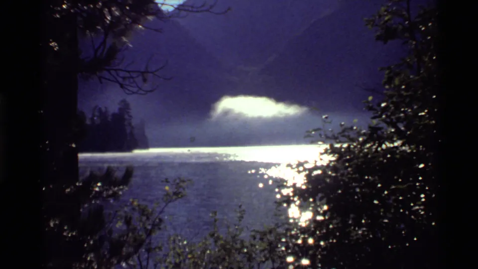1972 Holland Lake, Montana view framed by dark trees, with sunlit water and mist against a mountain backdrop.