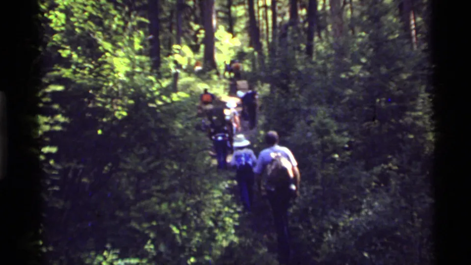 A small hiking group continues up the wooded Holland Lake trail, seen from behind in grainy 8mm footage.