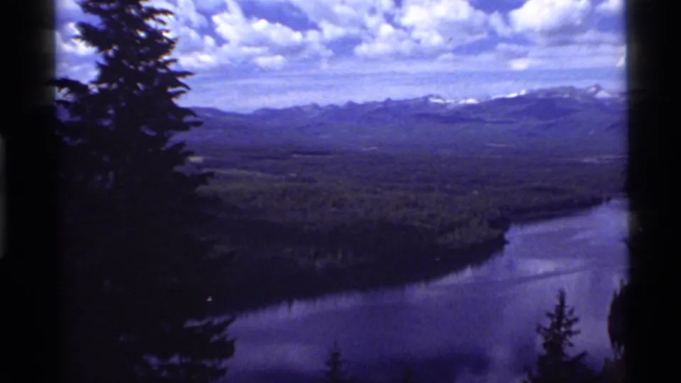 Scenic view of Holland Lake, Montana with forested shoreline, distant mountains, and partly cloudy sky in 1972 8mm footage.