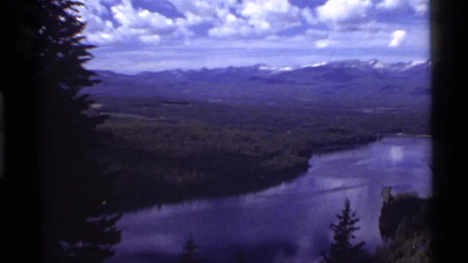 1972 8mm view of Holland Lake, Montana, seen from above with dark foreground trees, forested shoreline, and snow-capped mountains beyond.