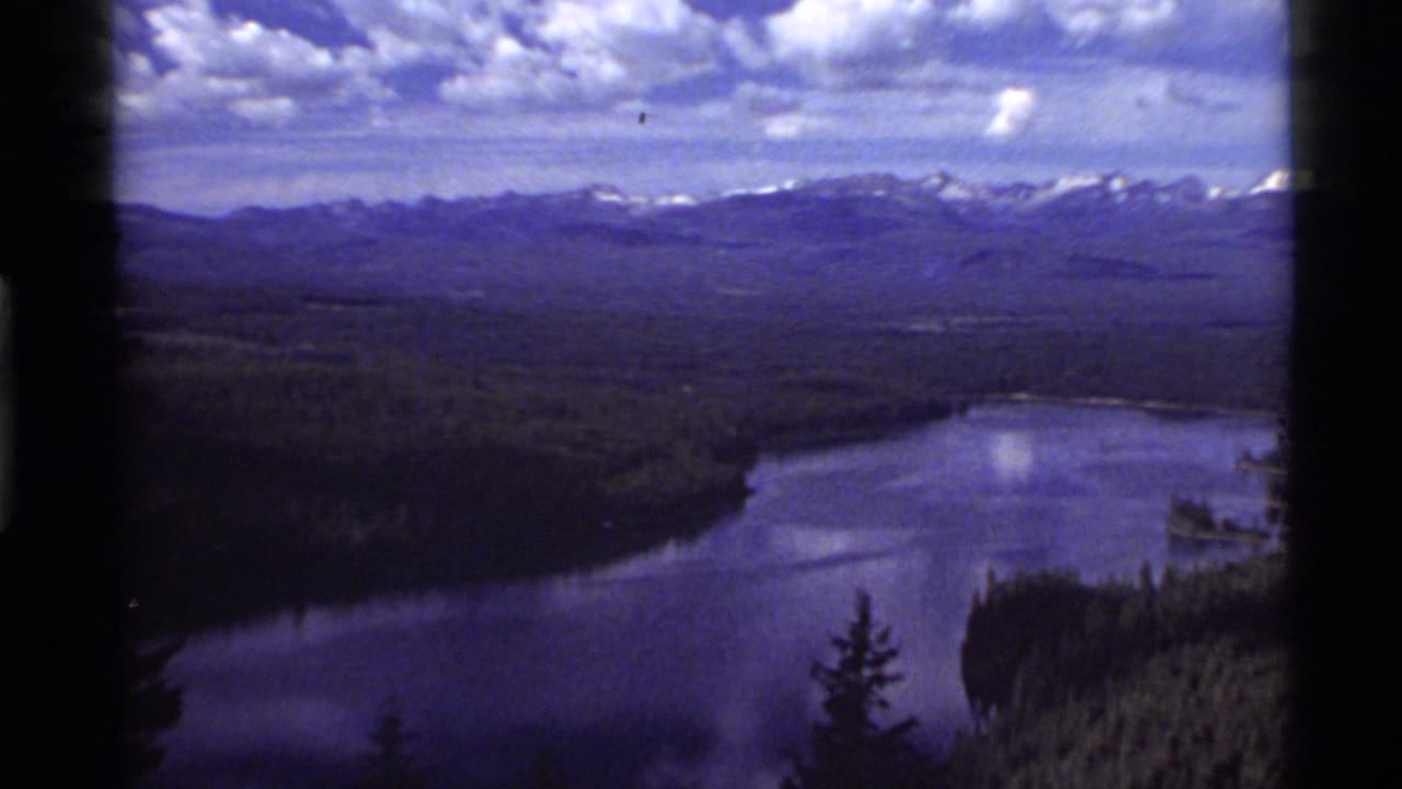 Holland Lake appears below a forested basin, with a purple-blue 8mm tint, distant Montana mountains, and a cloud-filled sky.