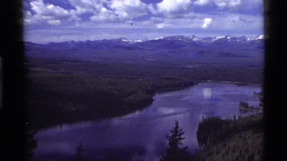 Holland Lake appears below a forested basin, with a purple-blue 8mm tint, distant Montana mountains, and a cloud-filled sky.