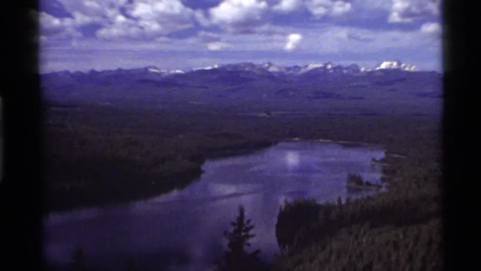 A wider 1972 home-movie frame shows Holland Lake bordered by dense forest, with snow-capped ridges and textured clouds across the horizon.