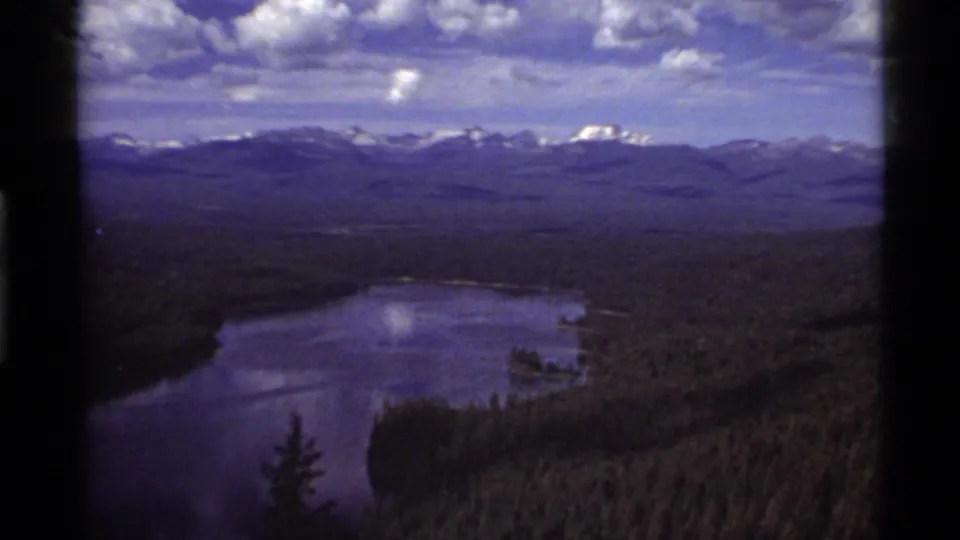 1972 8mm view over Holland Lake in Montana, with evergreen forest, distant snowy mountains, and dark film-edge borders.