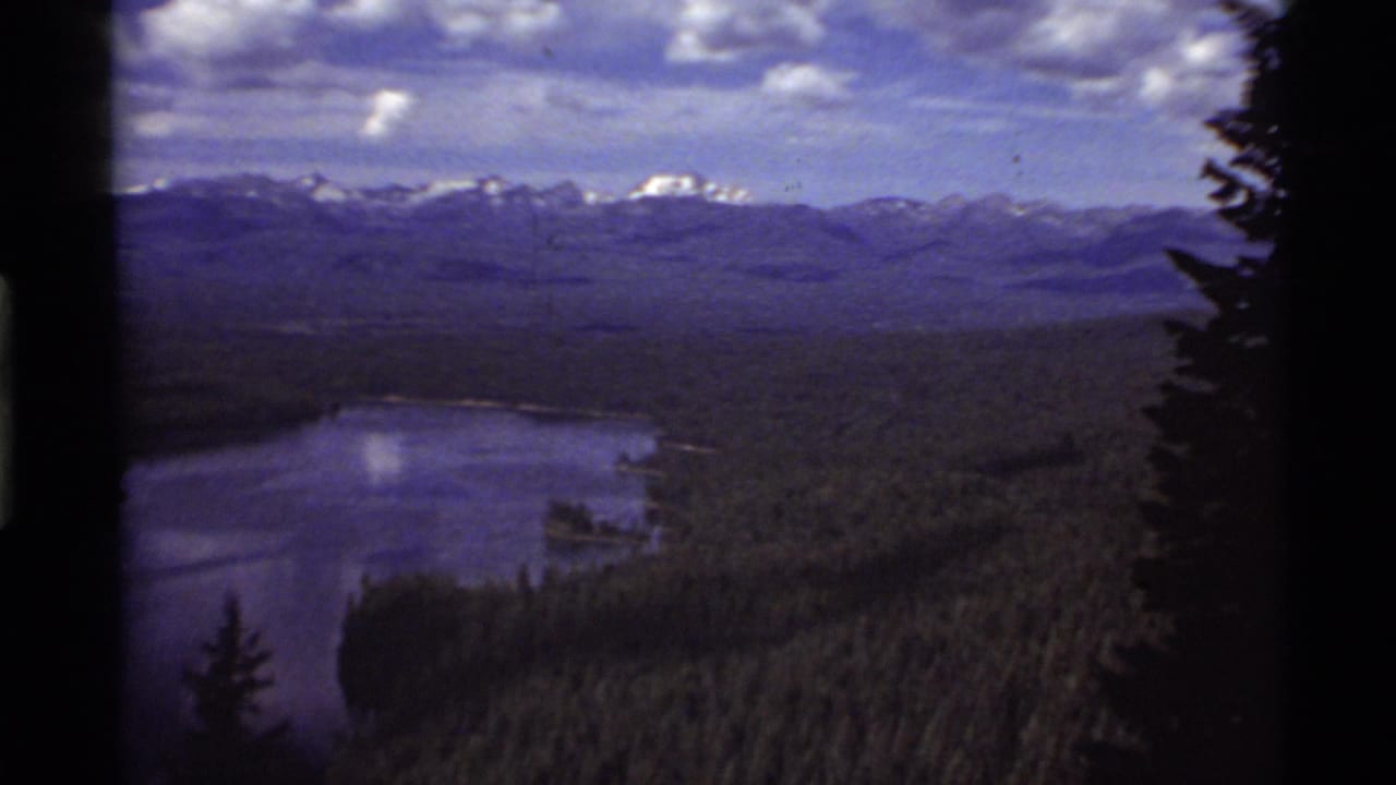 Lake and forest landscape with distant mountains, cloud cover, and a dark vertical film edge at center.