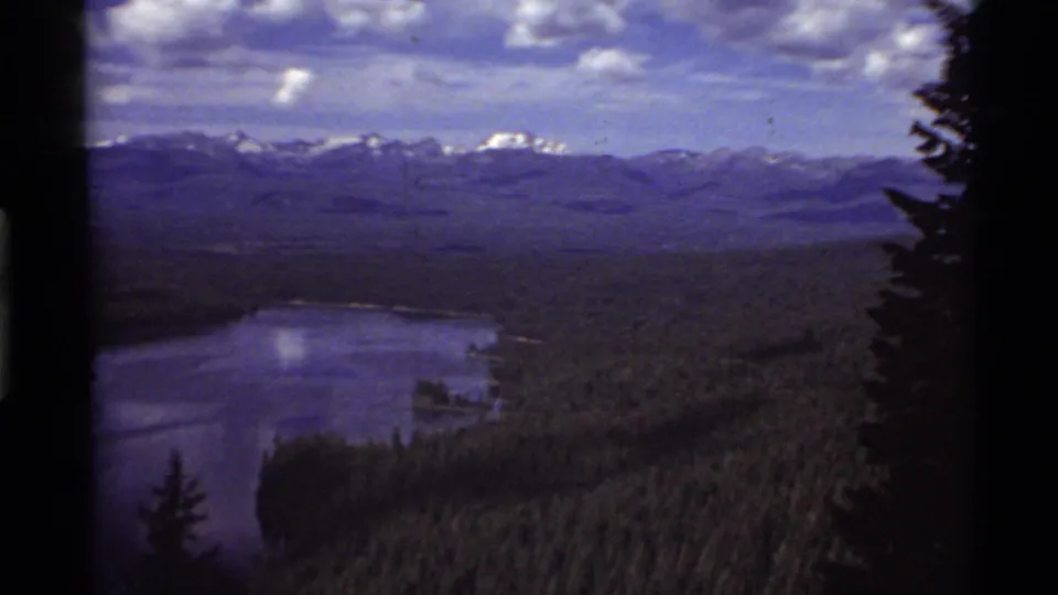 Panoramic 8mm frame of Holland Lake, Montana, showing blue water, evergreen shoreline, distant mountain range, and visible film border shadows.