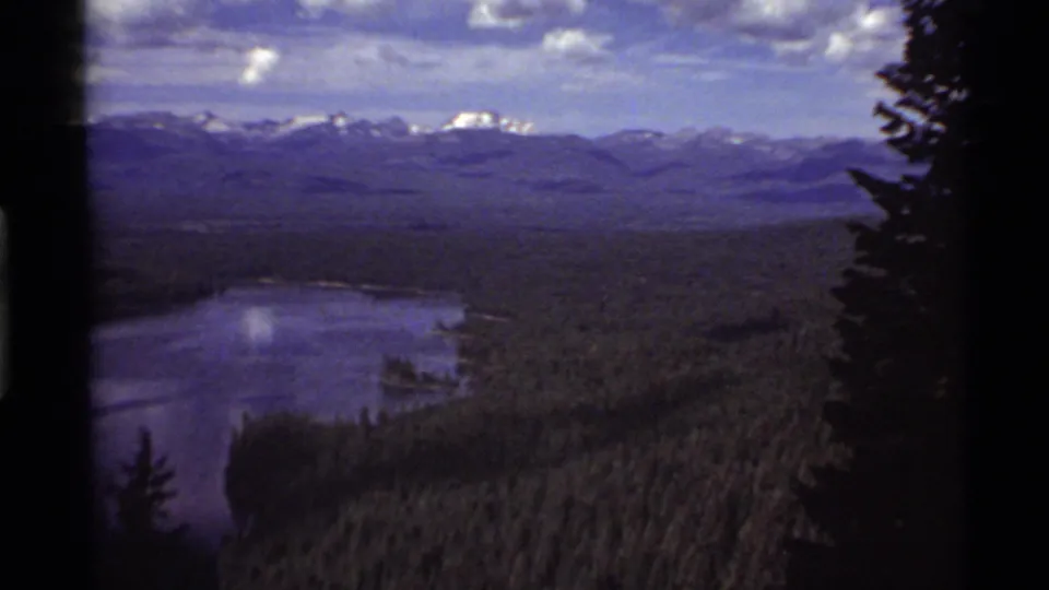 1972 home-movie frame from Holland Lake, Montana, with a wooded basin, lake surface, distant snowy peaks, and dark foreground tree forms.
