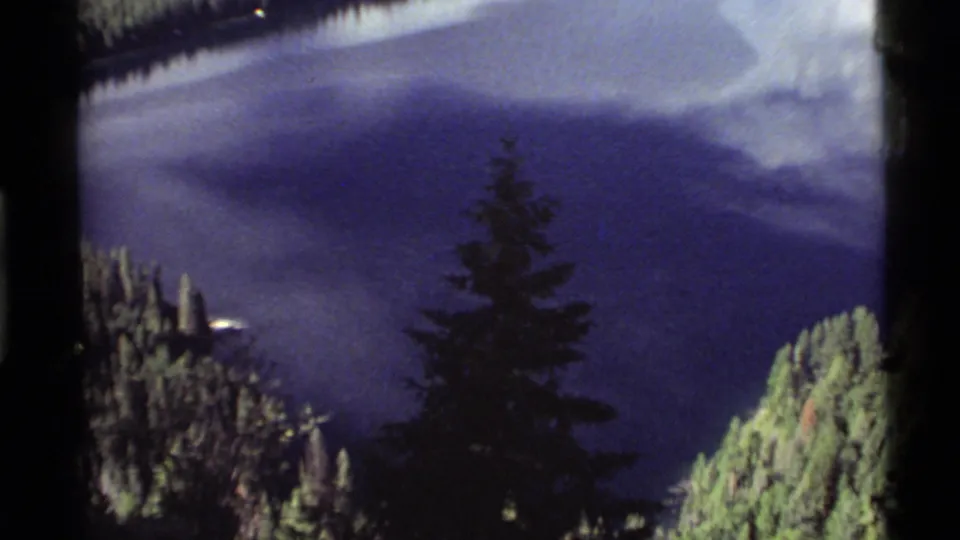 Later frame from the 1972 Holland Lake 8mm clip, showing deep blue water, green forested slopes, and a foreground evergreen silhouette.