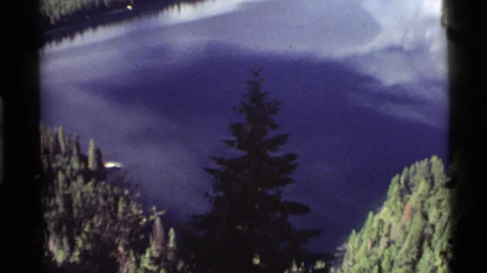 1972 8mm view at Holland Lake, Montana, showing blue water, forested shore, and a dark pine reflection in soft focus.
