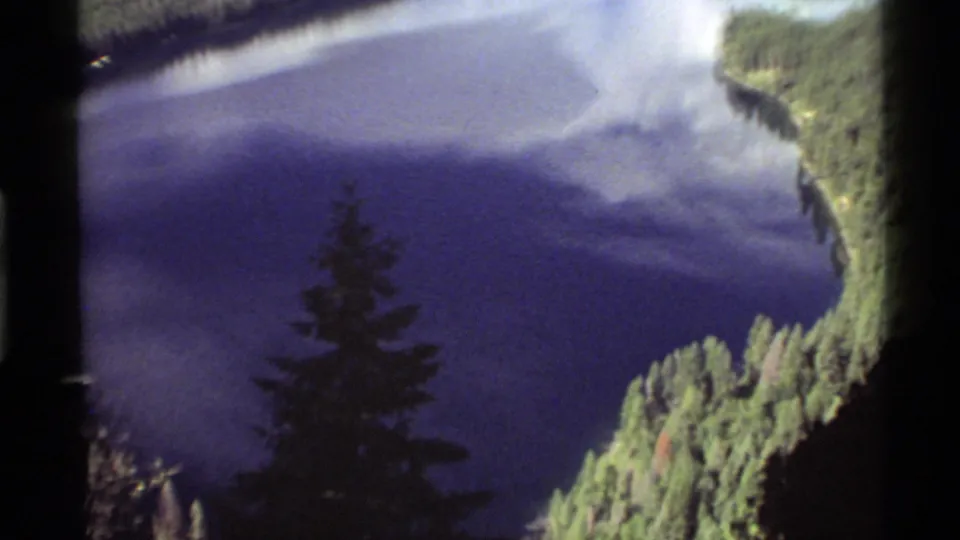 Soft 8mm frame of Holland Lake with evergreen banks, reflected sky on the water, and a silhouetted pine shape.