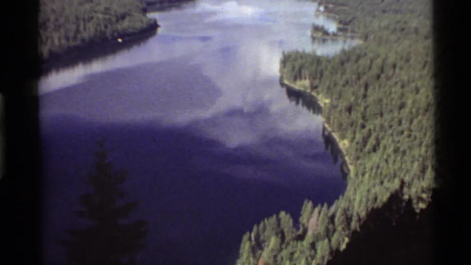 1972 Holland Lake frame showing a wider stretch of blue water, forested shoreline, sky reflection, and dark foreground tree silhouette.