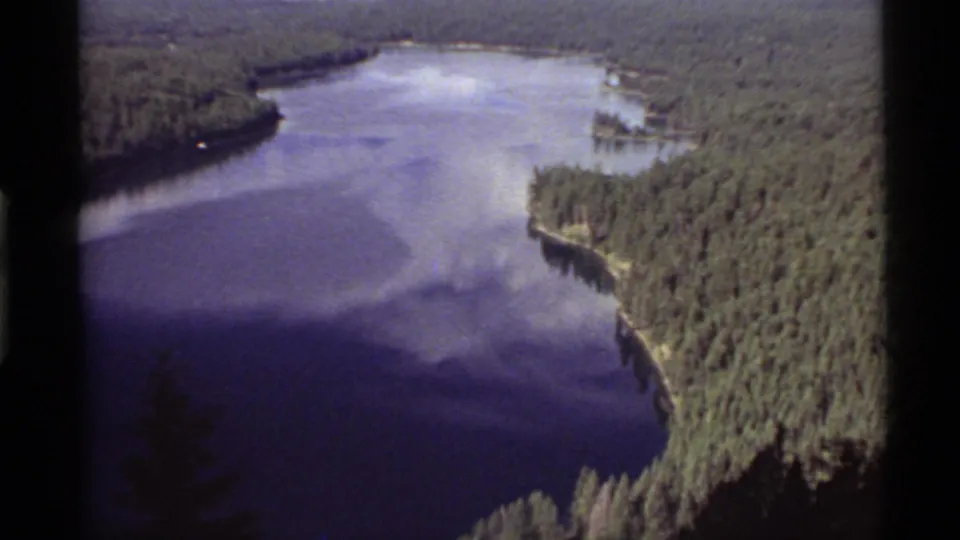 Vintage 8mm view of Holland Lake, Montana, with a forested bend, blue water, cloud reflections, and dark frame-edge shadows.