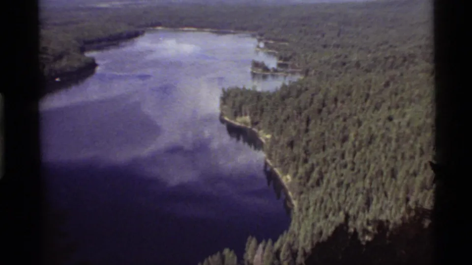 1972 8mm aerial view of Holland Lake, Montana, with blue water, forested shoreline, and a tree-covered peninsula.